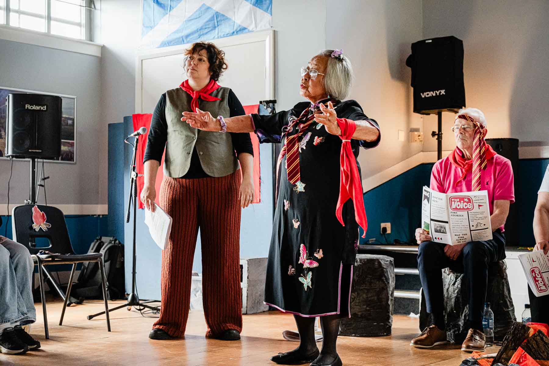 Two performers gesture expressively mid-scene, one in a green waistcoat, the other in a black dress with butterfly appliqués and a red neckerchief, with an audience member reading "The Voice" newspaper prop nearby.