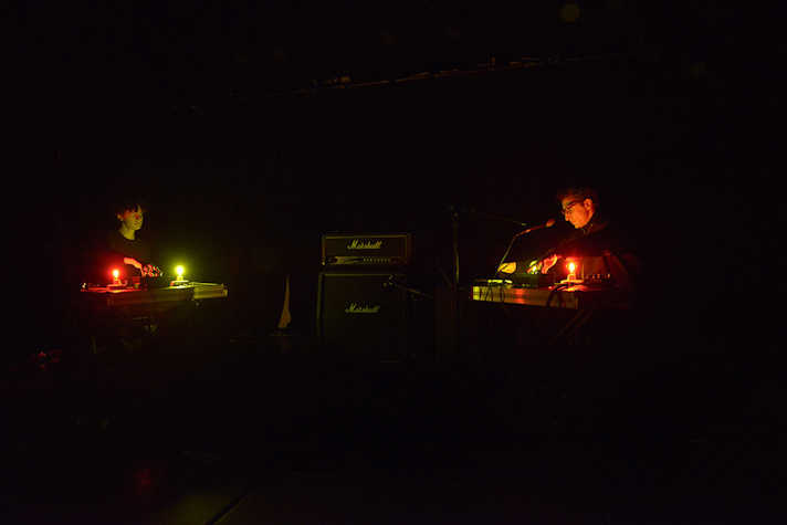 A dark photograph with a two people dimly lit, they are sitting at two desks lit by orange and green lights.