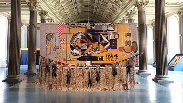 An image of Nirbhai (Nep) Singh Sidhu's installation in the Gallery of Modern Art, Glasgow. The image shows a large scale, colourful tapestry hung from the ceiling. There are tassels trailing on the floor. 