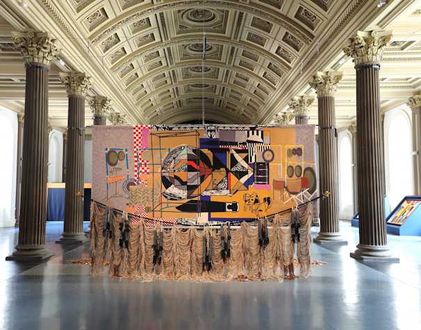An image of Nirbhai (Nep) Singh Sidhu's installation in the Gallery of Modern Art, Glasgow. The image shows a large scale, colourful tapestry hung from the ceiling. There are tassels trailing on the floor.