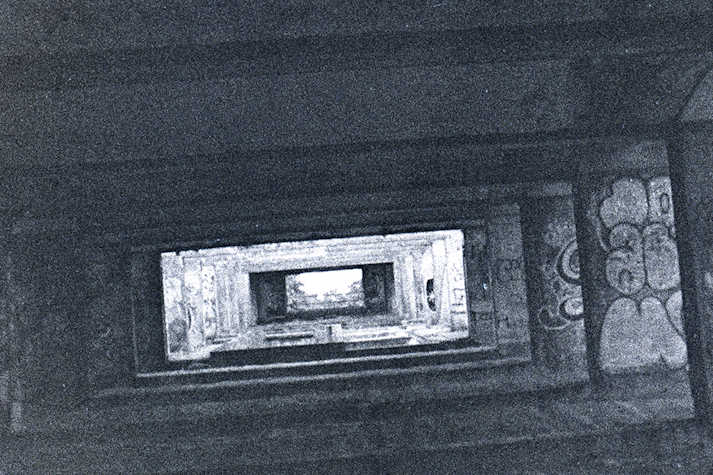 A photo with a grainy texture in black and white of showing a view through a concrete structure which is painted with graffiti. 