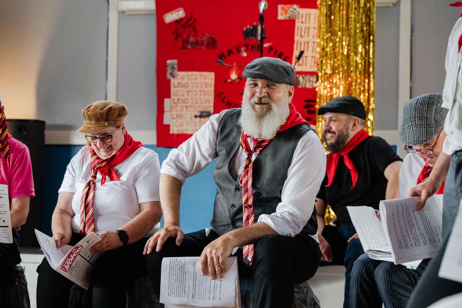 A performer in a purple jacket, bowler hat, and monocle reads from a script animatedly, with three other cast members behind her, in front of a projected screen and Saltire flag.