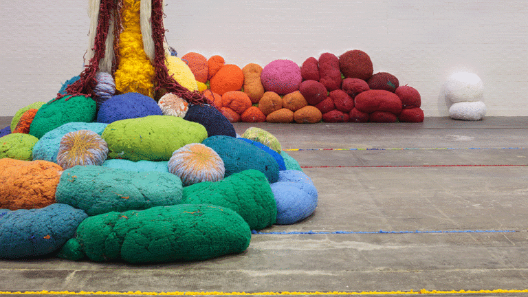 A view of coloured material forming soft sculptural shapes in a gallery. The materials are all different colours, and are shown in two sections, partly against a wall, and partly in a free-standing configuration