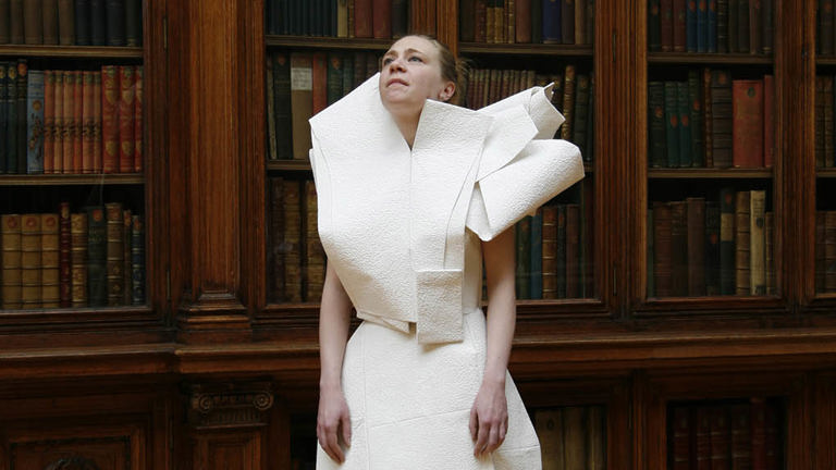 person stood looking upwards wearing a white dress that seems to be made out of paper, behind are dark bookshelves filled with books.