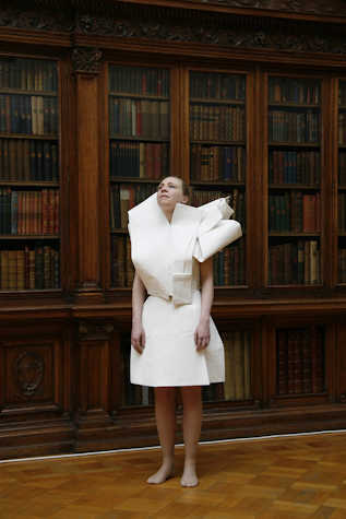 person stood looking upwards wearing a white dress that seems to be made out of paper, behind are dark bookshelves filled with books.