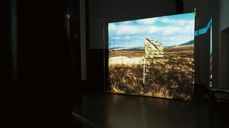 In a dark room a film is projected onto a screen. The project image shows a flag made from a gold emergency blanket, flying in an open field with hills in the far distance and a blue sky