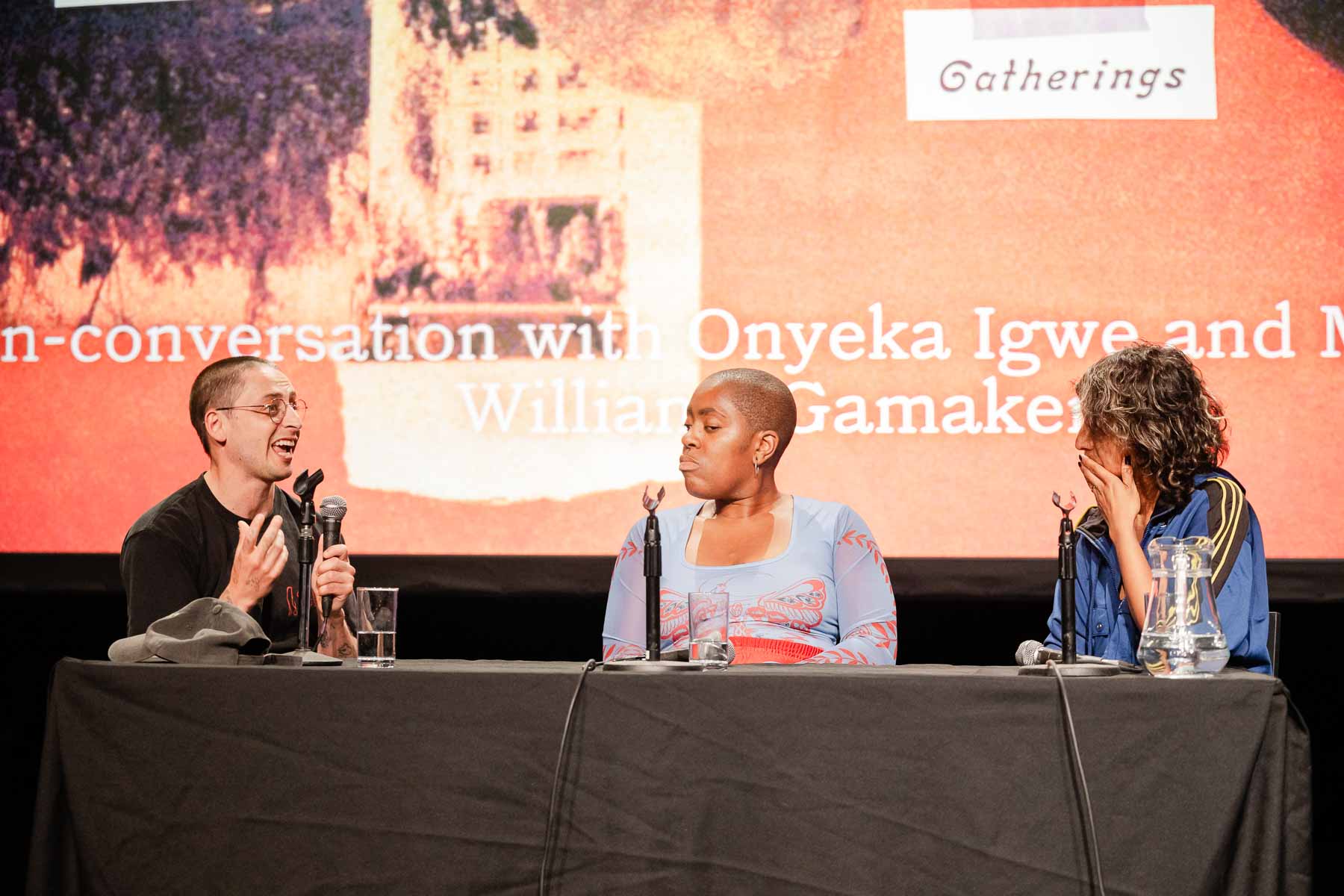 Three panellists in animated conversation at a table on stage, the man on the left speaking with an open expression, the woman in the centre reacting, and the woman on the right laughing with her hand to her face.
