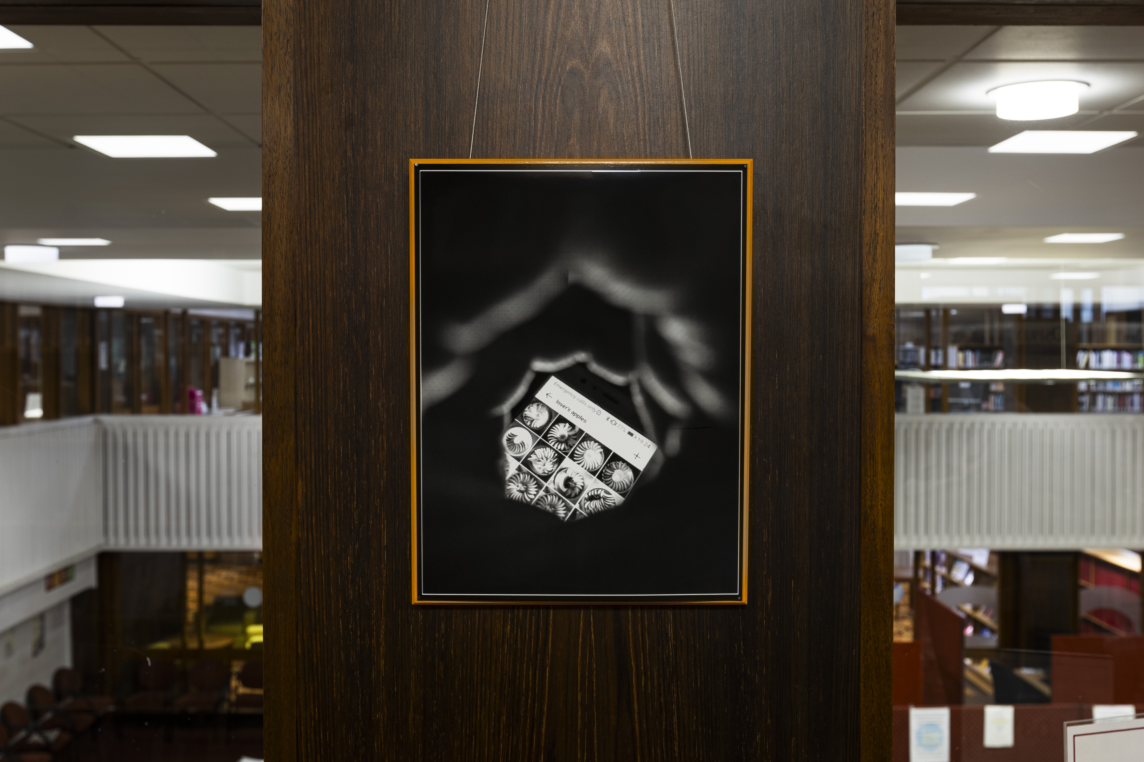 Orange-framed artwork on a wooden pillar showing cupped hands holding a smartphone displaying a locked screen with a photo grid titled "lover's apples," against a dark background.