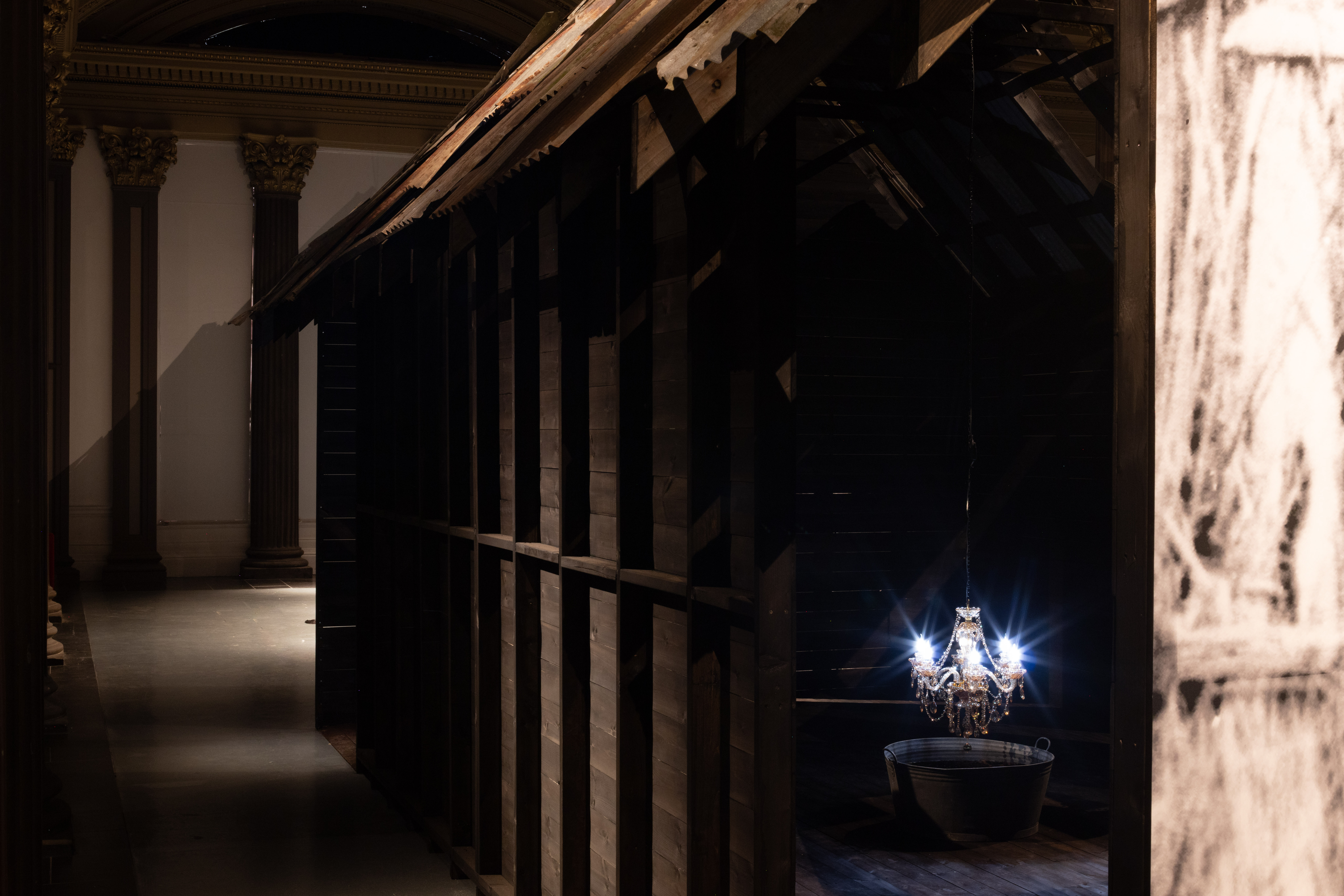 Installation view of a dark timber-framed structure built inside a grand columned gallery, with a lit crystal chandelier suspended over a metal wash tub visible through the wooden slats.