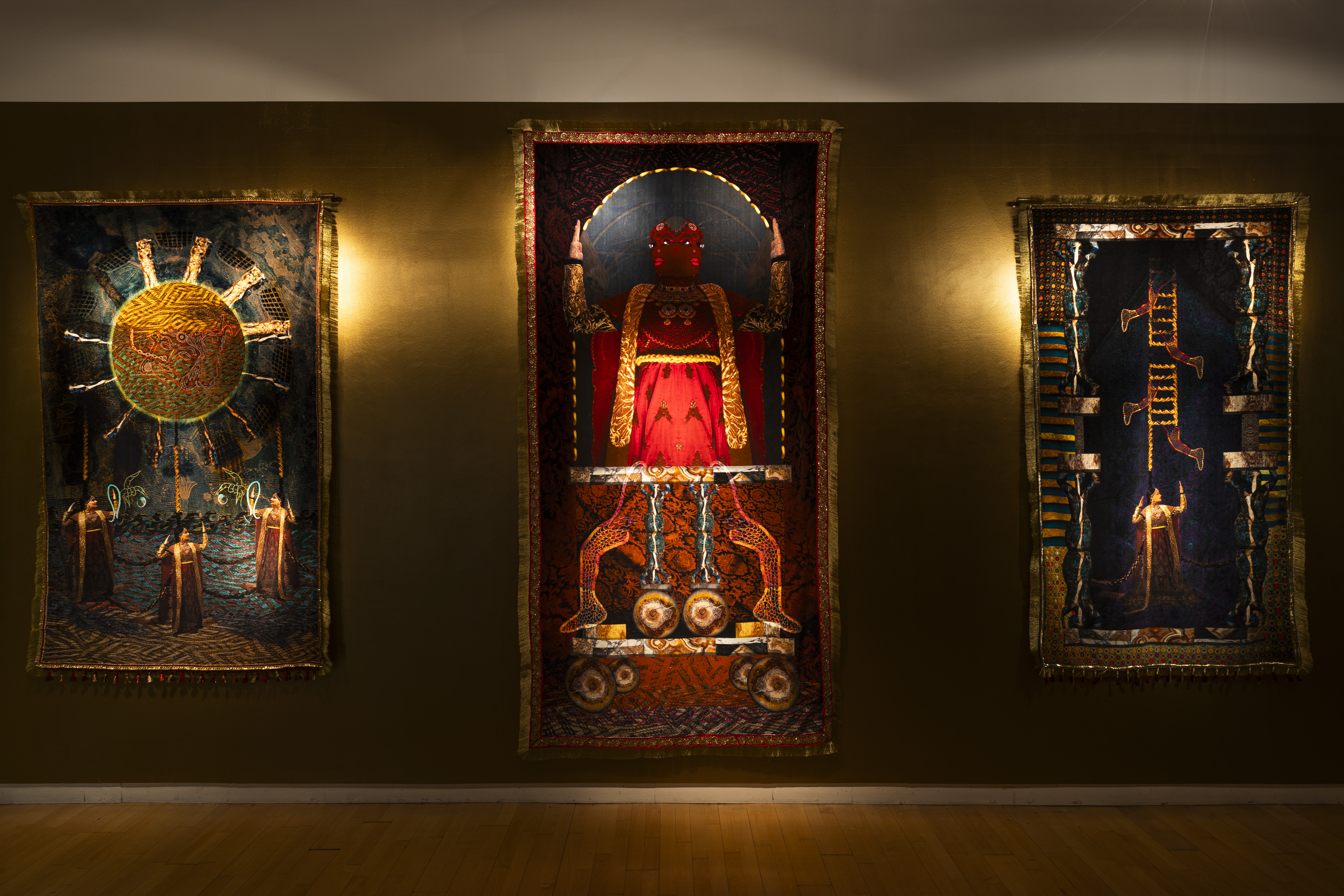 Three ornate tapestries by Aqsa Arif hung on a golden-brown wall at Streetlevel Photoworks, depicting a radiant sun with robed figures, a red-cloaked deity-like figure on a throne, and a ladder-like structure with falling figures.