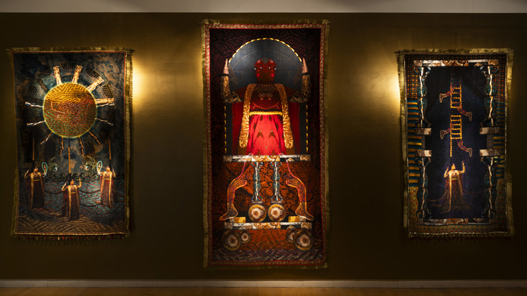 Three ornate tapestries by Aqsa Arif hung on a golden-brown wall at Streetlevel Photoworks, depicting a radiant sun with robed figures, a red-cloaked deity-like figure on a throne, and a ladder-like structure with falling figures.