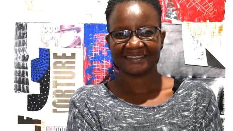 A woman stands against a wall which has collages against it, with her arms crossed, she is wearing a grey top.
