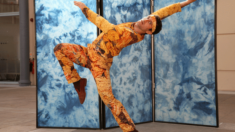 man stands on one leg, leaning back, moving expressively wearing a suit patterned with images of pasta in front of a trifold screen with blue marble patterning.