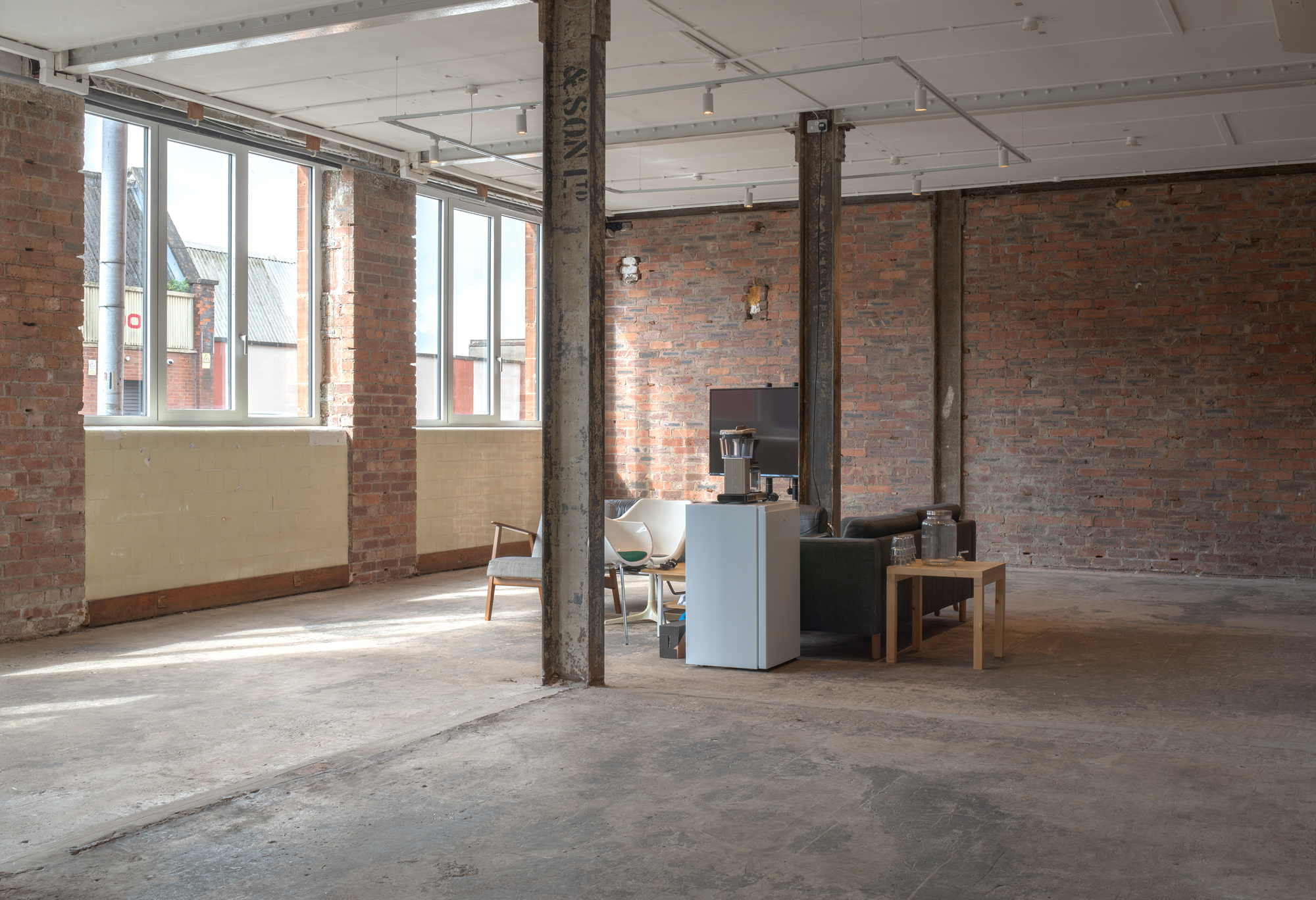 A large open-plan industrial space with exposed brick walls, steel columns and tall windows, furnished sparsely with a television, mismatched armchairs and a small side table.