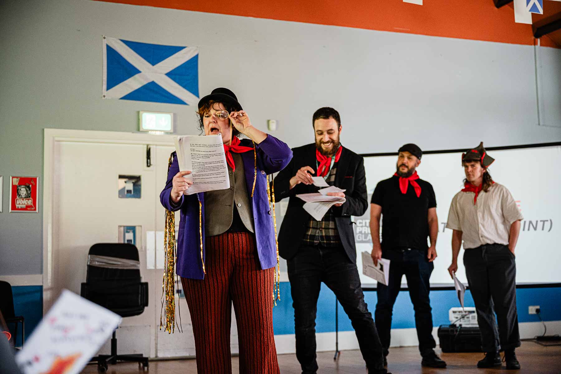 A performer in a purple jacket, bowler hat, and monocle reads from a script animatedly, with three other cast members behind her, in front of a projected screen and Saltire flag.