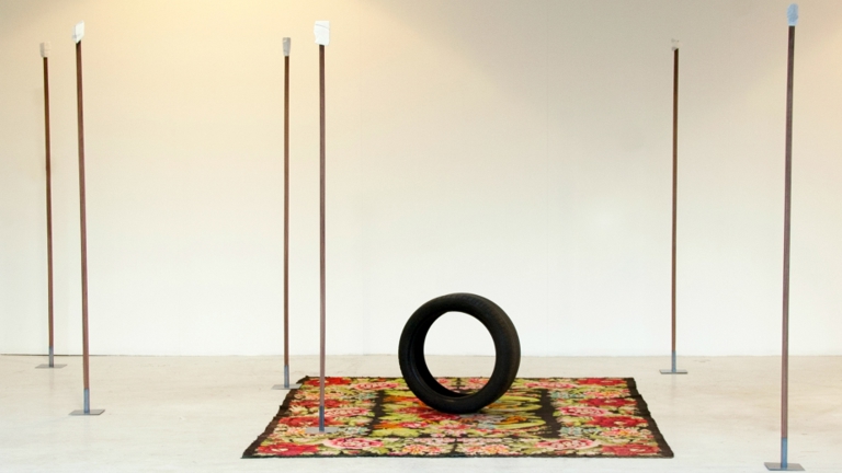 A white room with sculptural objects including a patterned rug with a car tyre on it. There are a number of vertical poles dotted around, with metal plates at the top of them 