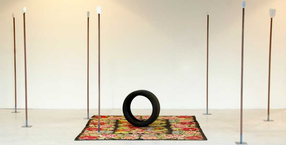 A white room with sculptural objects including a patterned rug with a car tyre on it. There are a number of vertical poles dotted around, with metal plates at the top of them