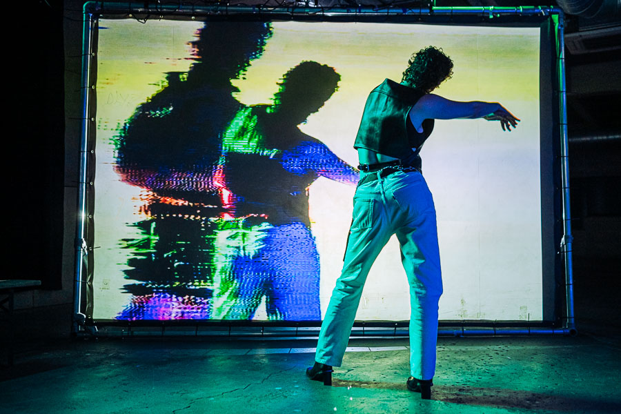 A performer in a leather waistcoat and cream trousers reaches an arm outward, seen from behind, against a large screen showing a glitched, colourful projection of two figures.