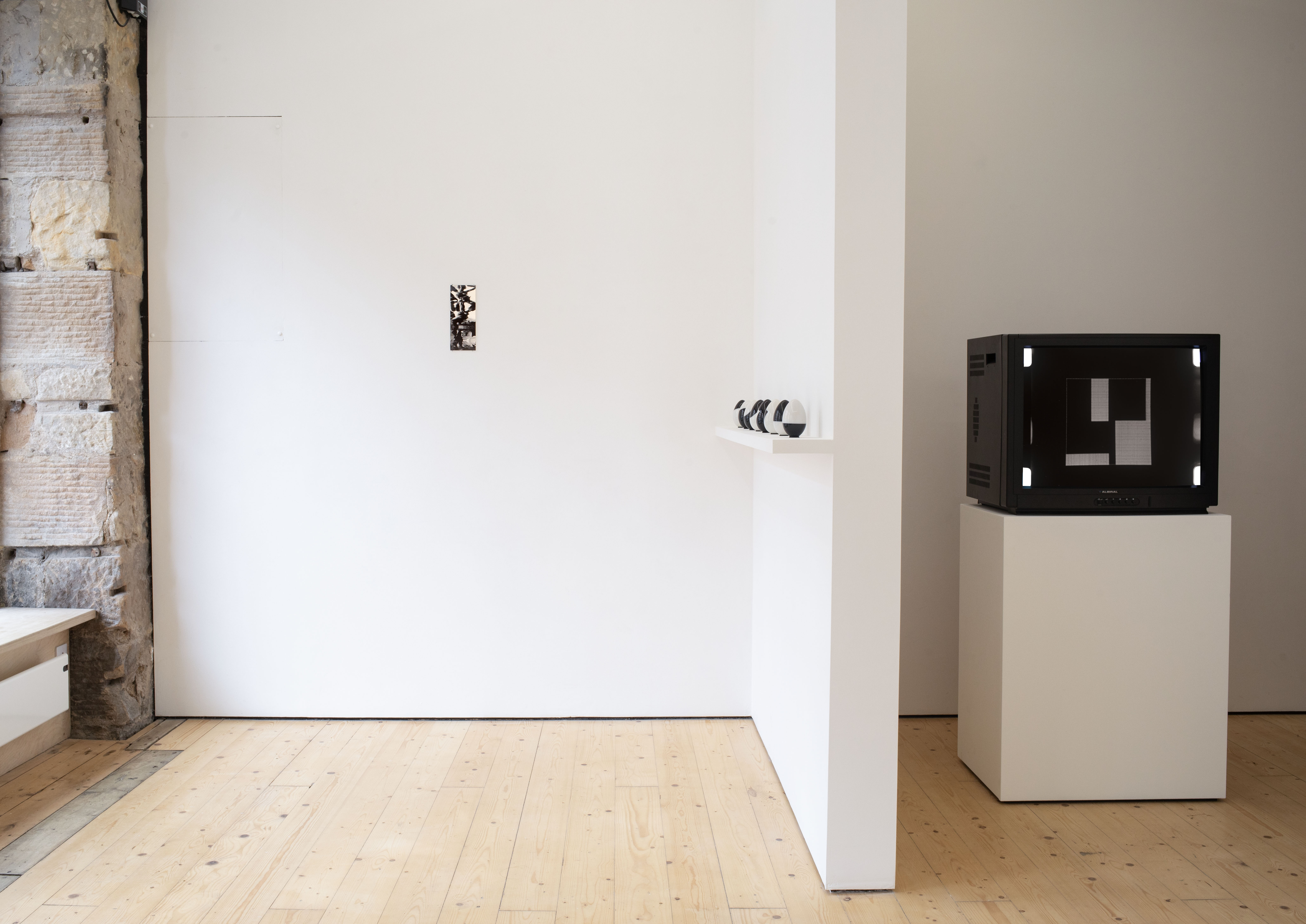 Gallery installation with a small black-and-white photograph on a white wall, a row of small black-and-white egg-shaped objects on a shelf, and a vintage television displaying a geometric pattern, set on a plinth beside exposed stone.