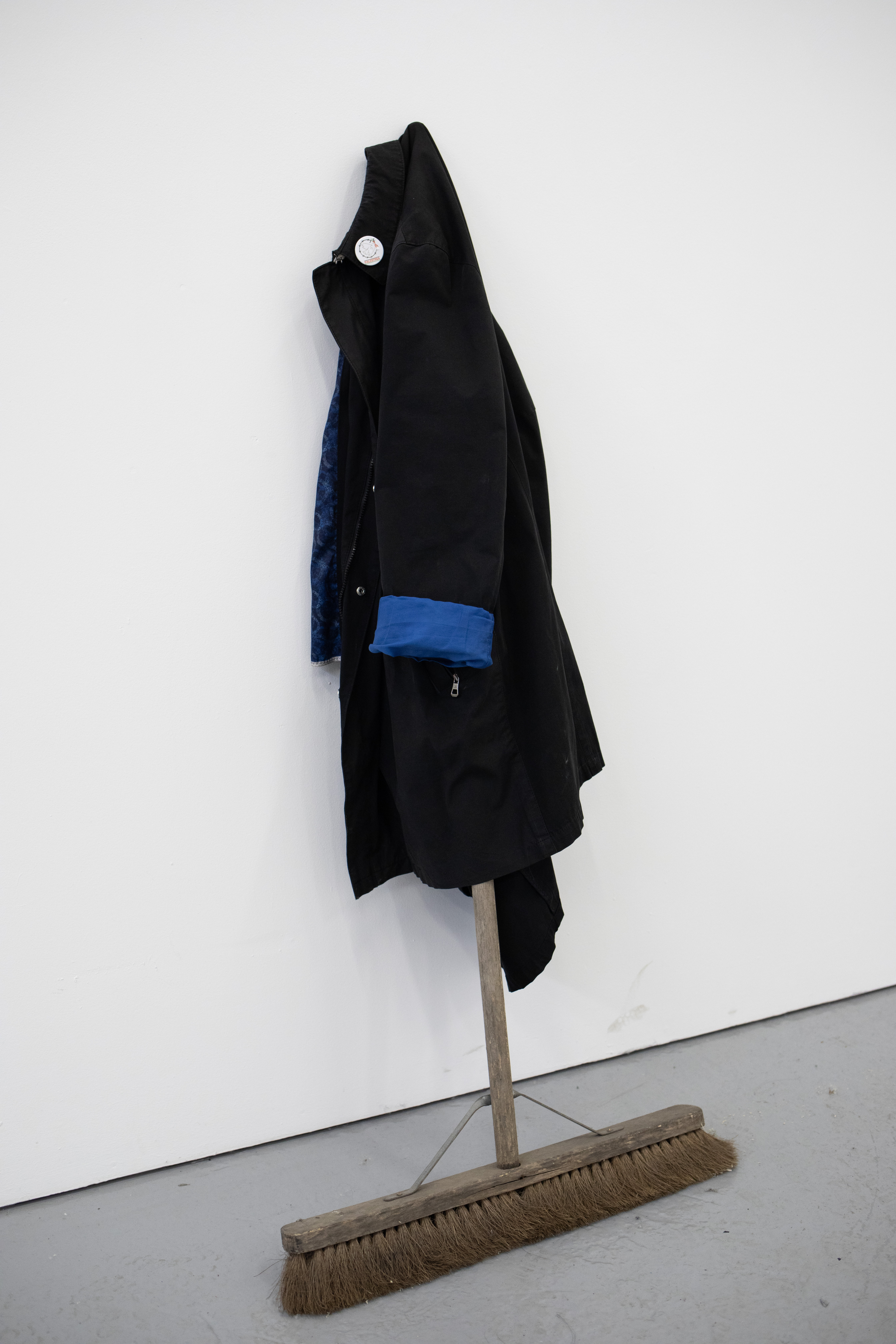 Sculptural installation of a black coat with a blue lining and cuff, hung on a broom propped against a white gallery wall, its bristles resting on the floor.