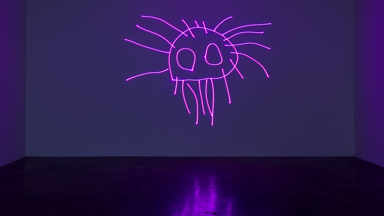A dark room with a purple hue, projected in the centre is a neon purple childrens drawing. The drawing features a circular shape with eyes and several legs. 