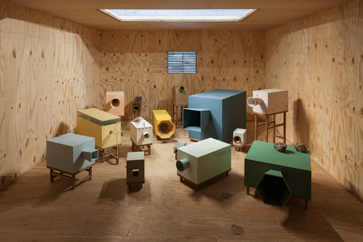 In a wooden room, there are multiple cubic sculptures, all in different colours. There is a vent in the centre of the back wall. The sculptures are all placed on wooden stands. Each of them has a different shape mouth opening, including circular, square and hexagon.