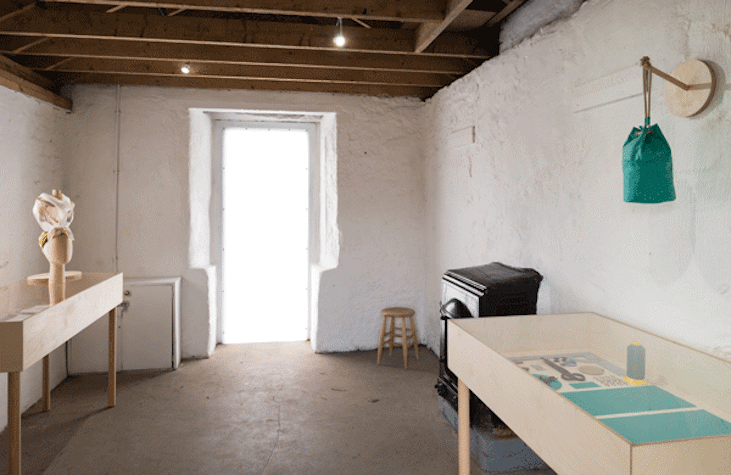 View of an exhibition space with an open door at the end. Vitrines are shown on either side, containing exhibition material. A stove is also visible.