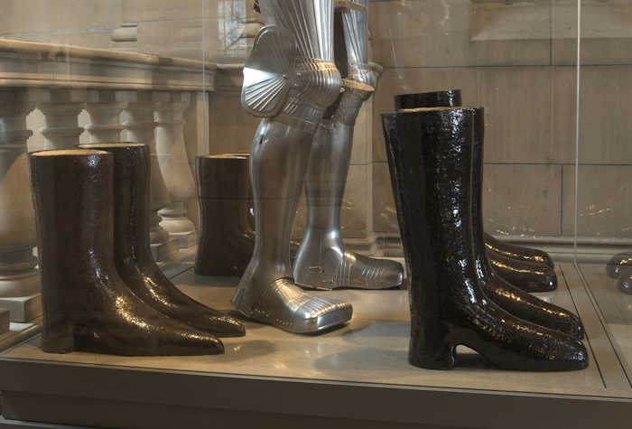 assortment of boots lined up around a knights armour boots, in a museum display case