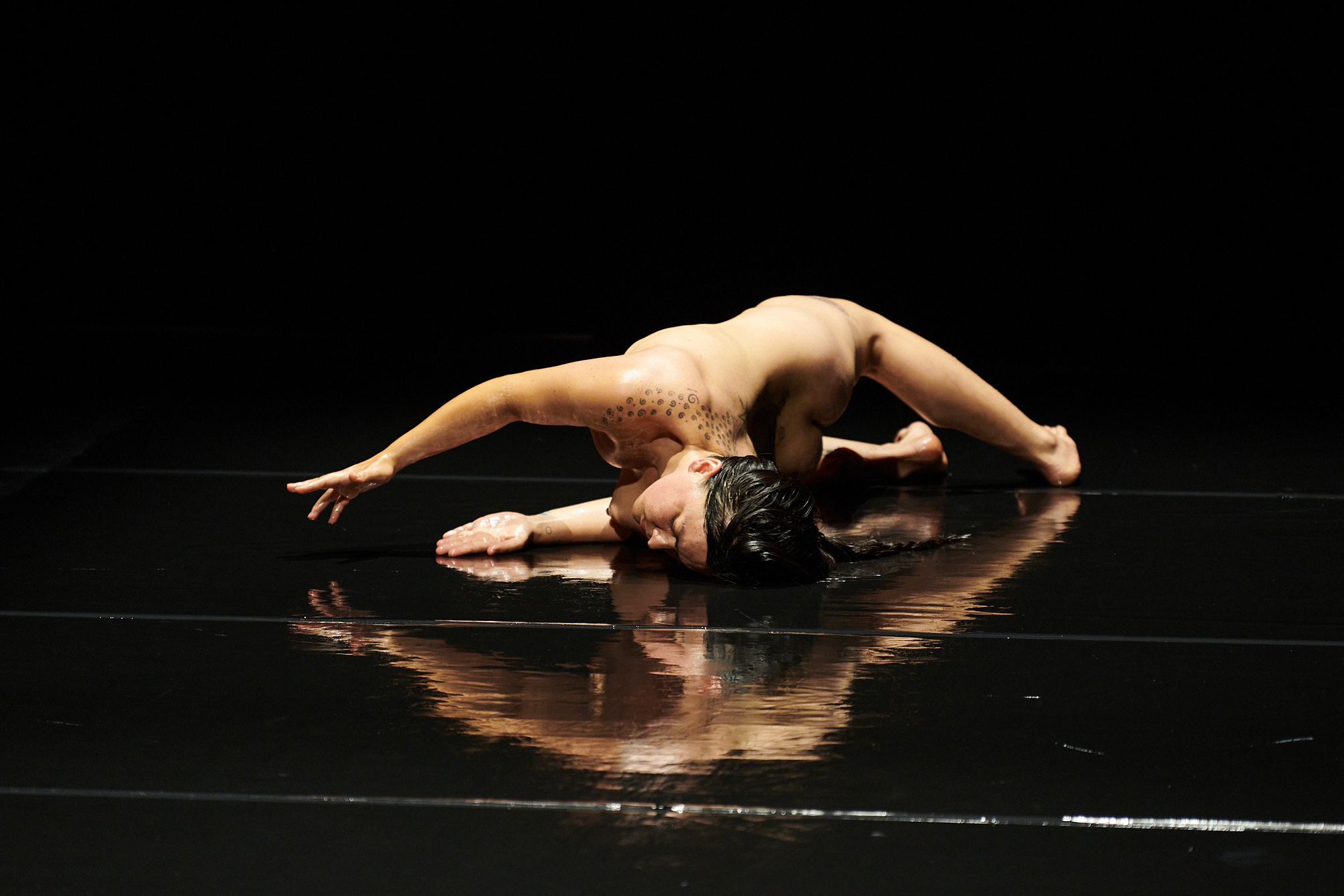  A nude woman with dark hair, olive skin and tattoos along her shoulder, lies on a reflective black surface, arches her back with her arms extended in front of her.   