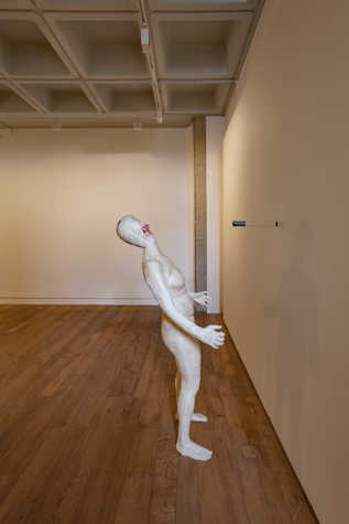 A white figurine leans back away from a wall, there is a bullet-like object in front of the face of the figurine.