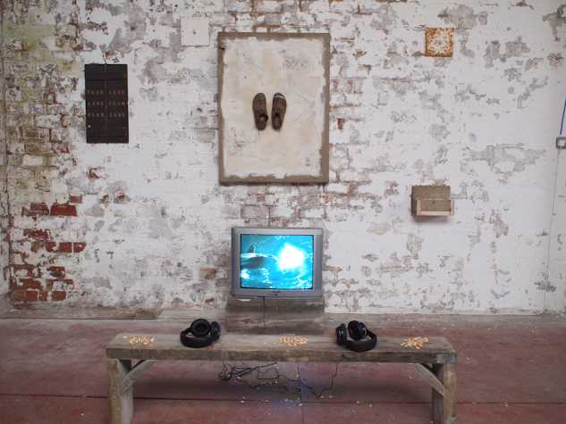 painted brick walled gallery space. in the centre hangs a canvas board with two brown shoes stuck onto the centre of it. around are three smaller pieces, one reads 'fear less less fear fear less'. beneath is a crt monitor showing water. in front a wooden bench with two pairs of headphones and nut shells.