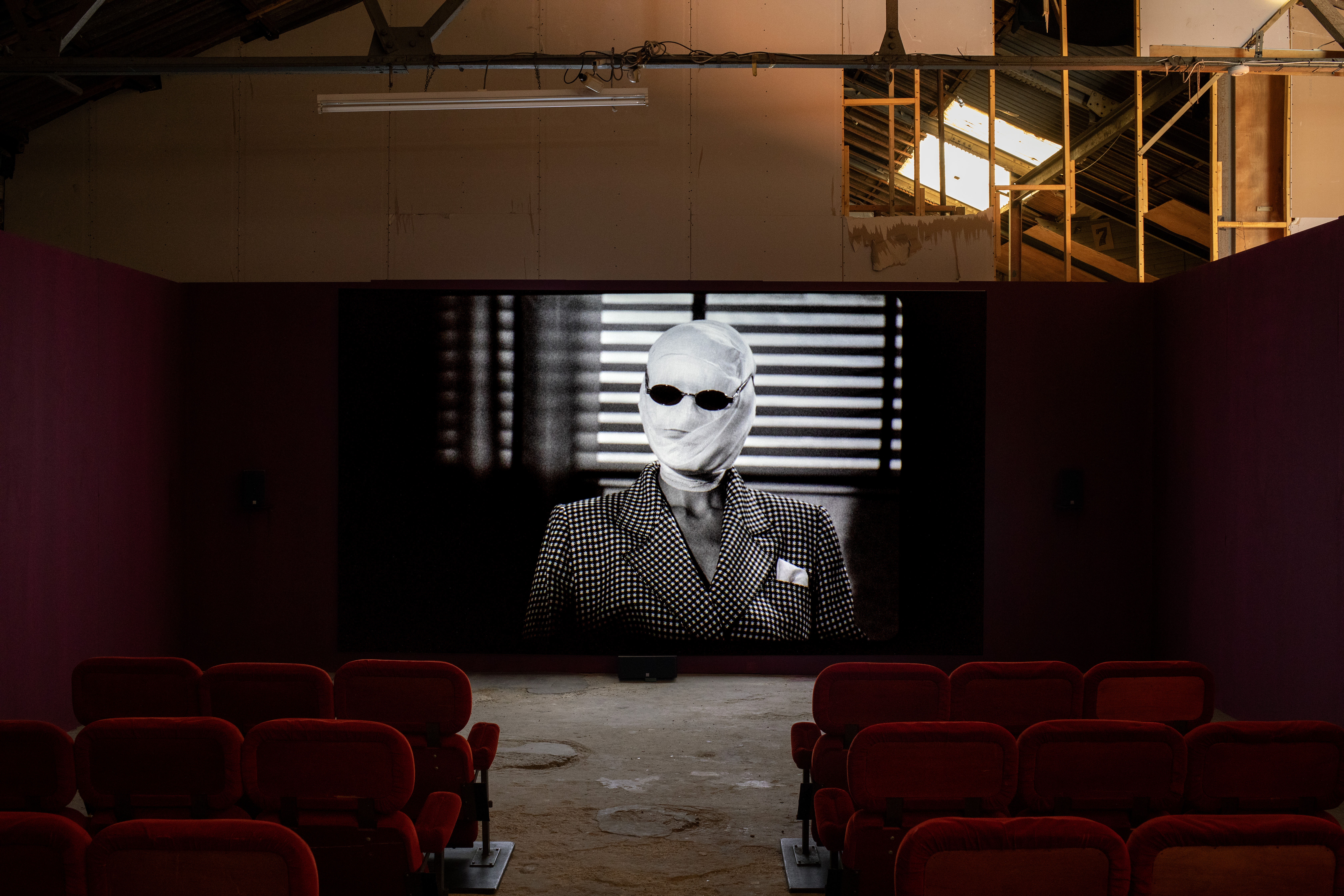 A large screen in a warehouse cinema space projecting a black and white still from Michelle Williams Gamaker's film "Strange Evidence," showing a figure with a bandaged head and dark sunglasses in a checked jacket, lit by striped window blinds, with rows of red velvet seating facing the screen.