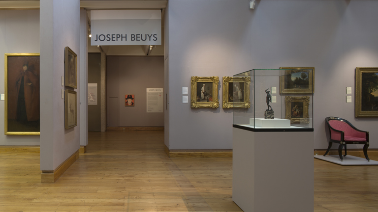 installation view of a gallery. on the wall it reads 'Joseph beuys'. surrounding there are  gilded framed works on a wall and a glass display case/plinth