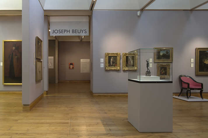 installation view of a gallery. on the wall it reads 'Joseph beuys'. surrounding there are  gilded framed works on a wall and a glass display case/plinth