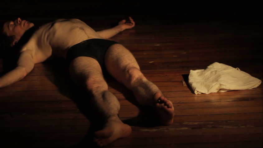 A white man has is sprawled on the wooden floor with only black underwear. A white cloth is to his right.