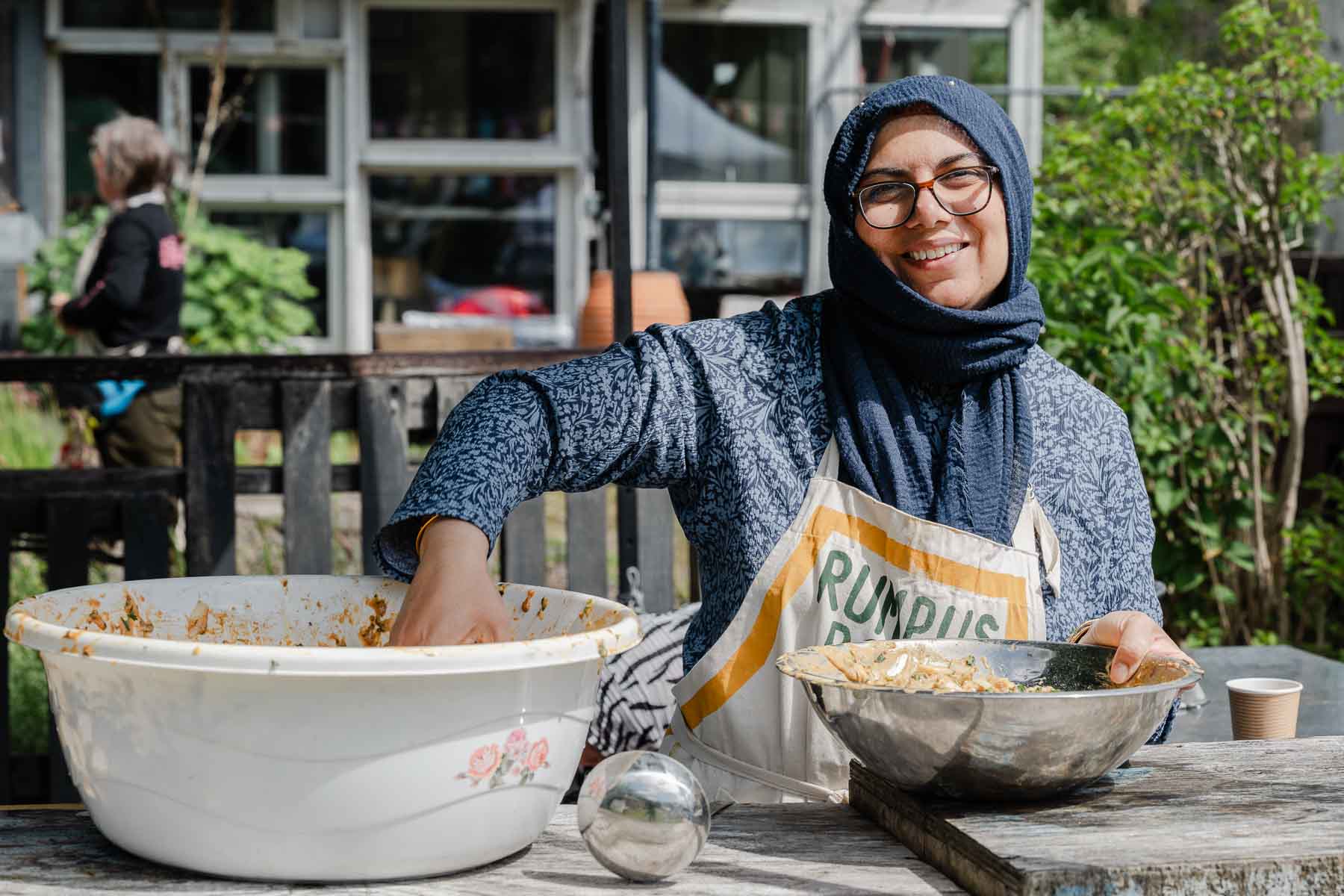 A smiling woman wearing a headscarf and a "Rumpus Room" apron mixes ingredients in a large metal bowl at an outdoor table, with washing-up bowls and a garden setting behind her.