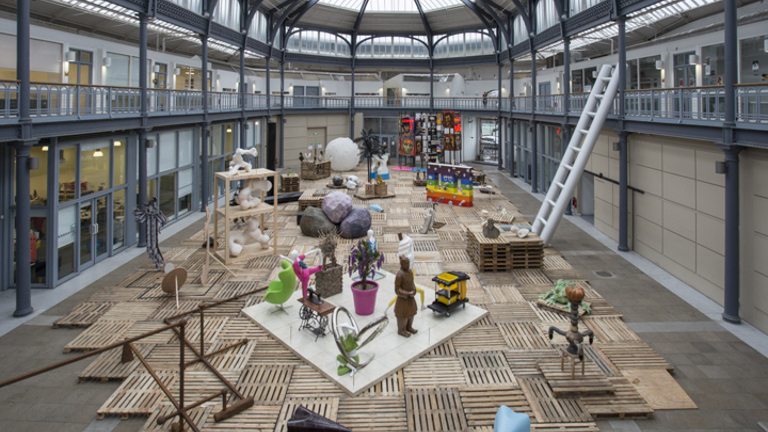 In a large hall, with a glass ceiling, in an aerial view, there are multiple sculptures placed on top of a temporary wooden flooring. 