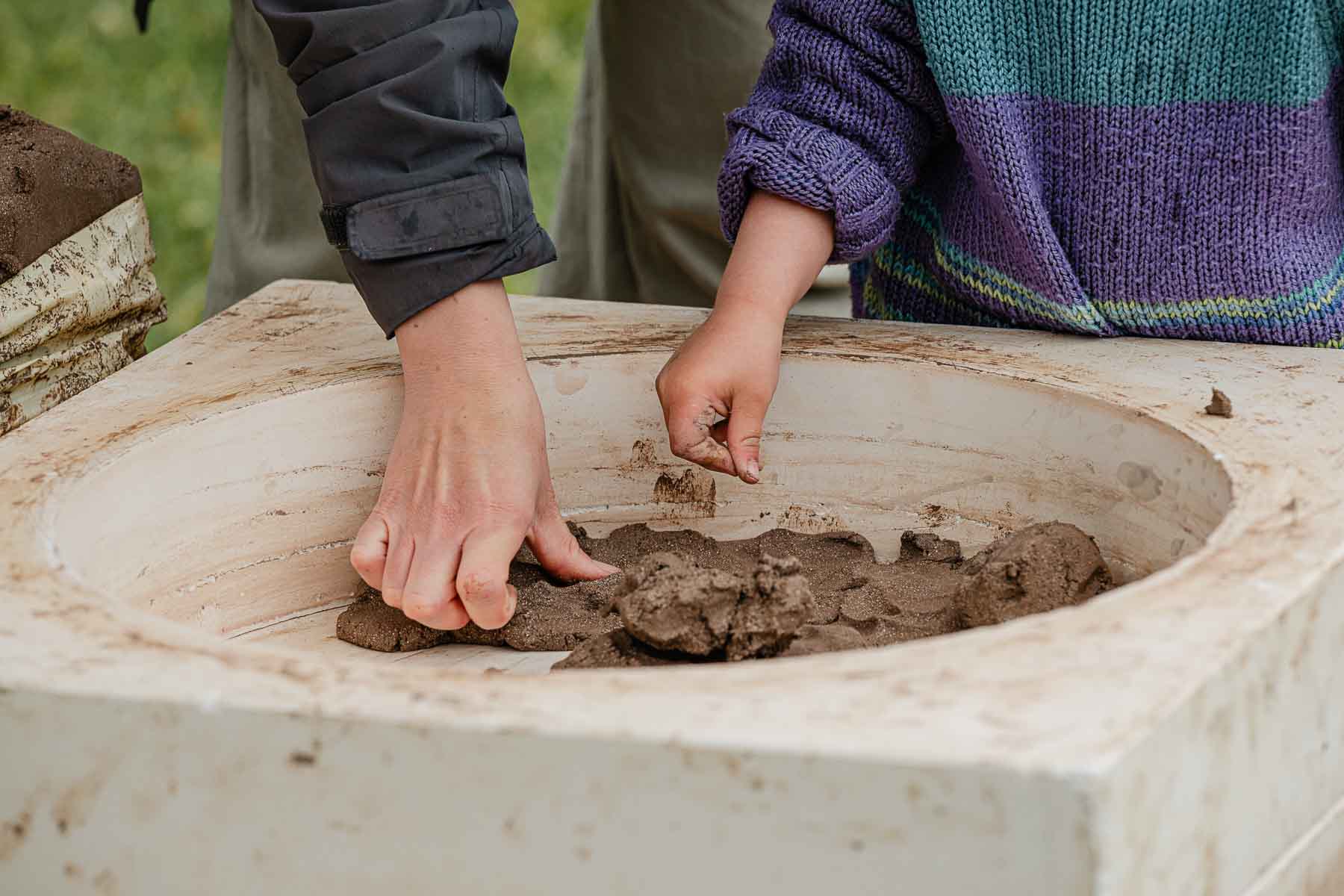 Close-up of two people's hands shaping wet clay in a large stone basin outdoors.