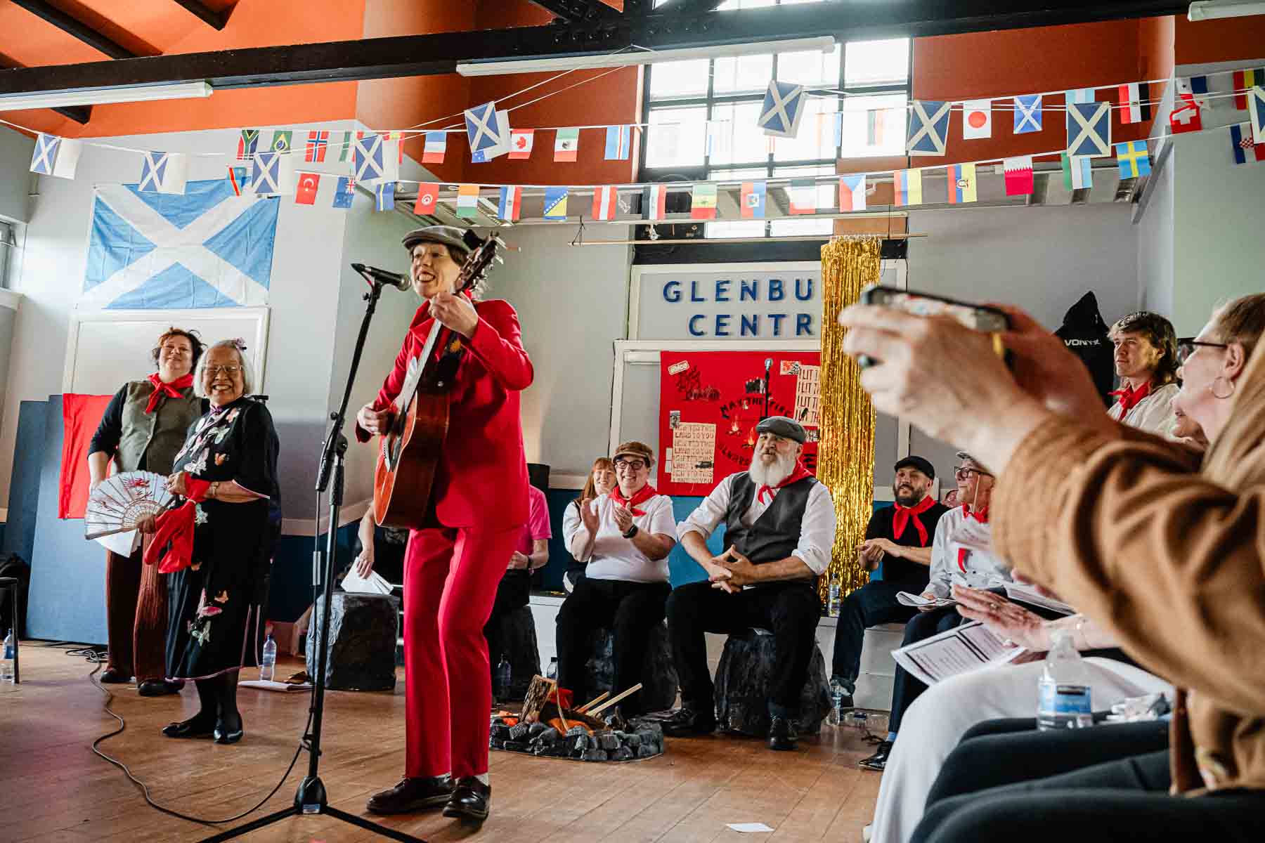 A performer in a red suit sings into a microphone while playing guitar, surrounded by seated cast members and an audience member filming on a phone, beneath flag bunting and a "Glenburn Centre" sign.