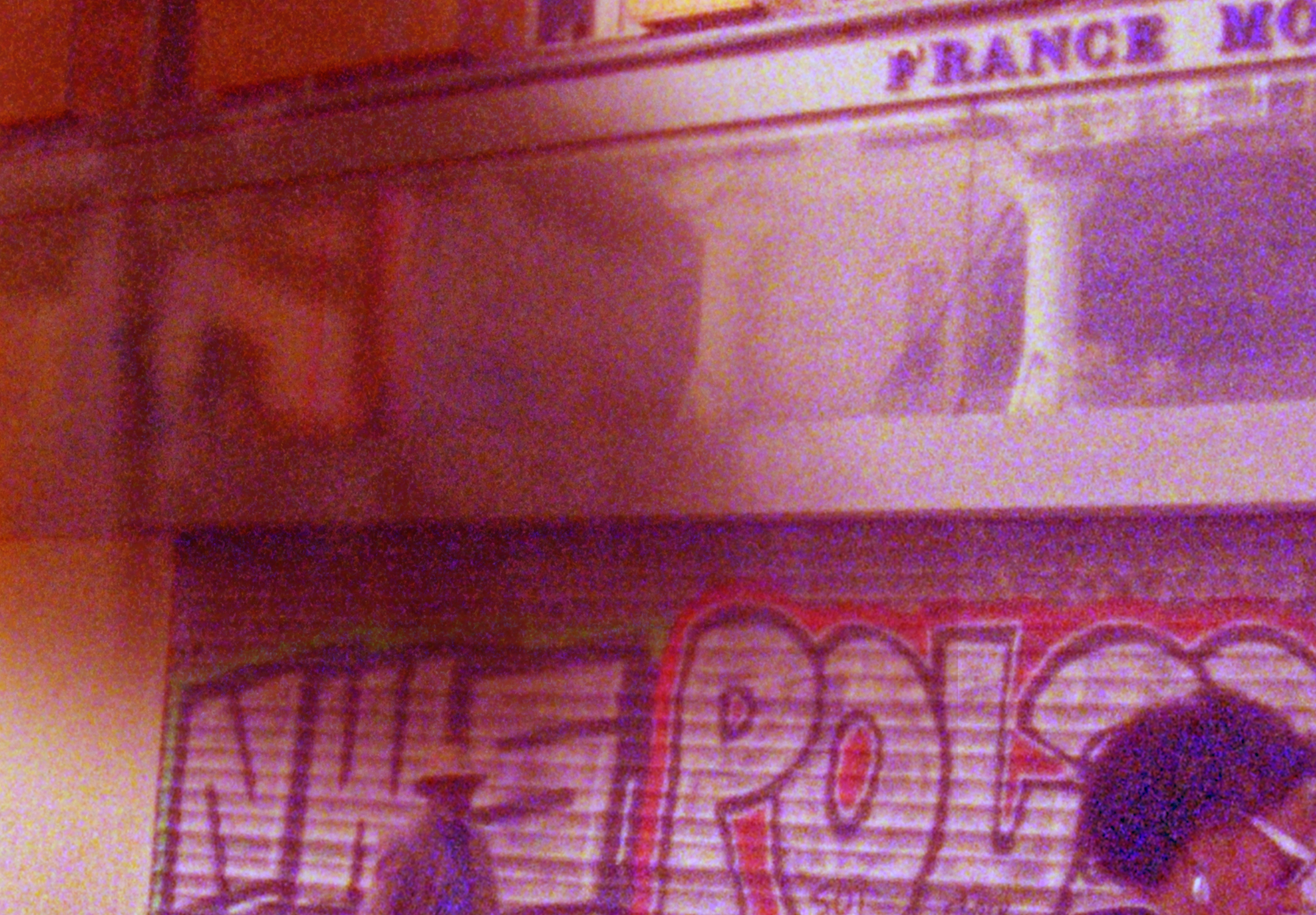 A grainy magenta-tones street image showing shopfronts with graffities shutters and people walking past. The image is hazy and blurred, with the word "FRANCE" clearly visibile on a building in the top right corner. 
