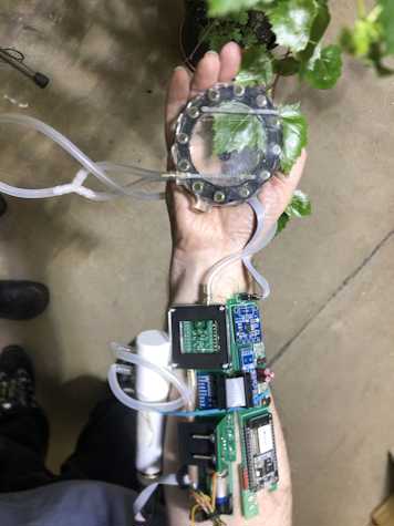 A photo of an outstretch arm and hand which is covered by a device with visible batteries, wiring and tubing. In their palm they hold a leaf below part of the machine.