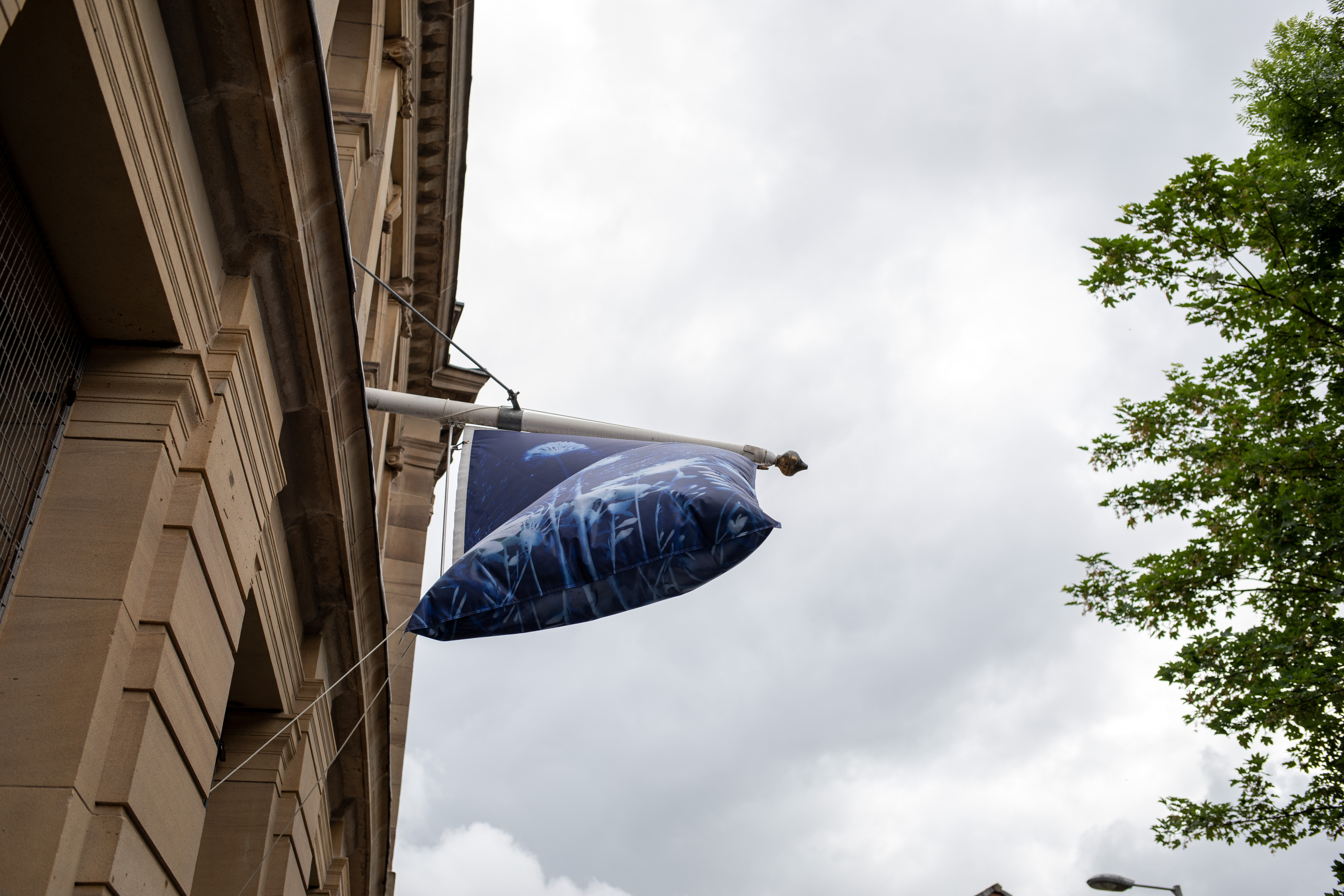 A cyanotype-printed flag, its fabric filled with wind, hangs from a stone building against a cloudy sky and green tree.
