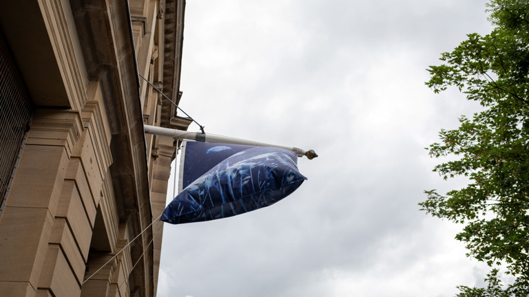 A cyanotype-printed flag, its fabric filled with wind, hangs from a stone building against a cloudy sky and green tree.