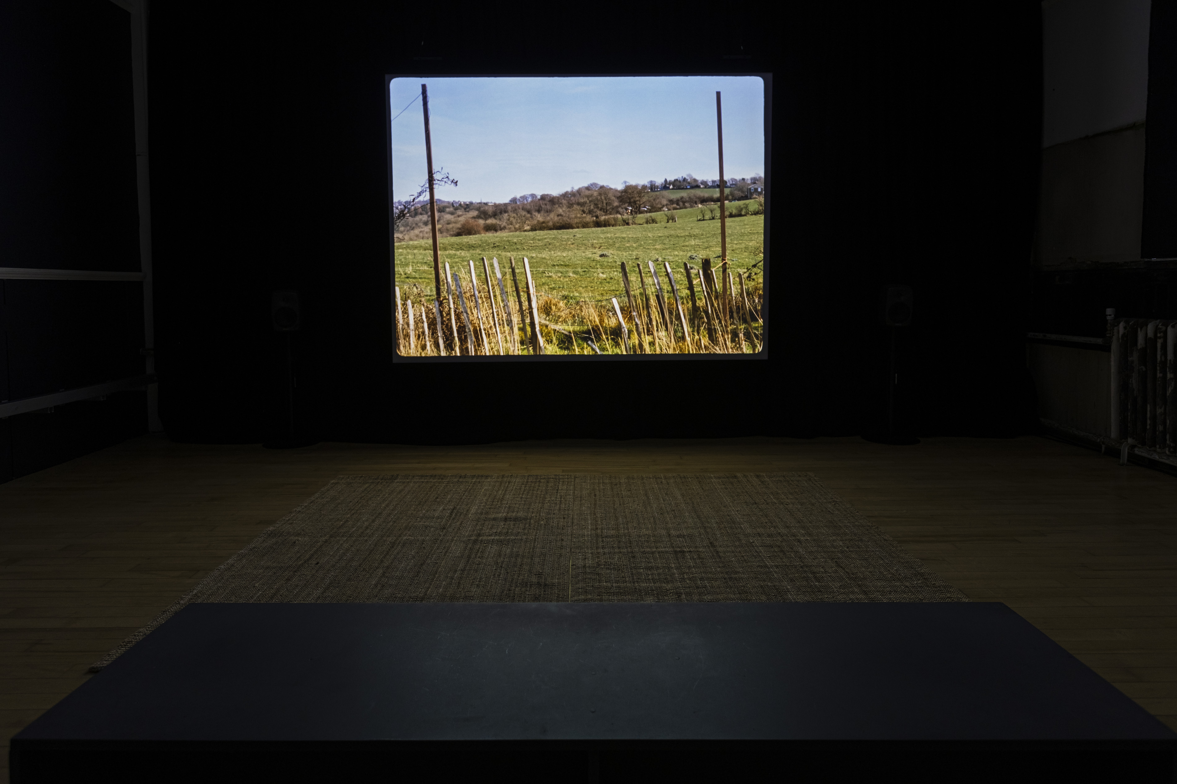 Darkened gallery installation with a large projected film still of a rural field with a wooden fence, viewed across a dark platform with a woven mat, part of a video work by Luke Fowler.