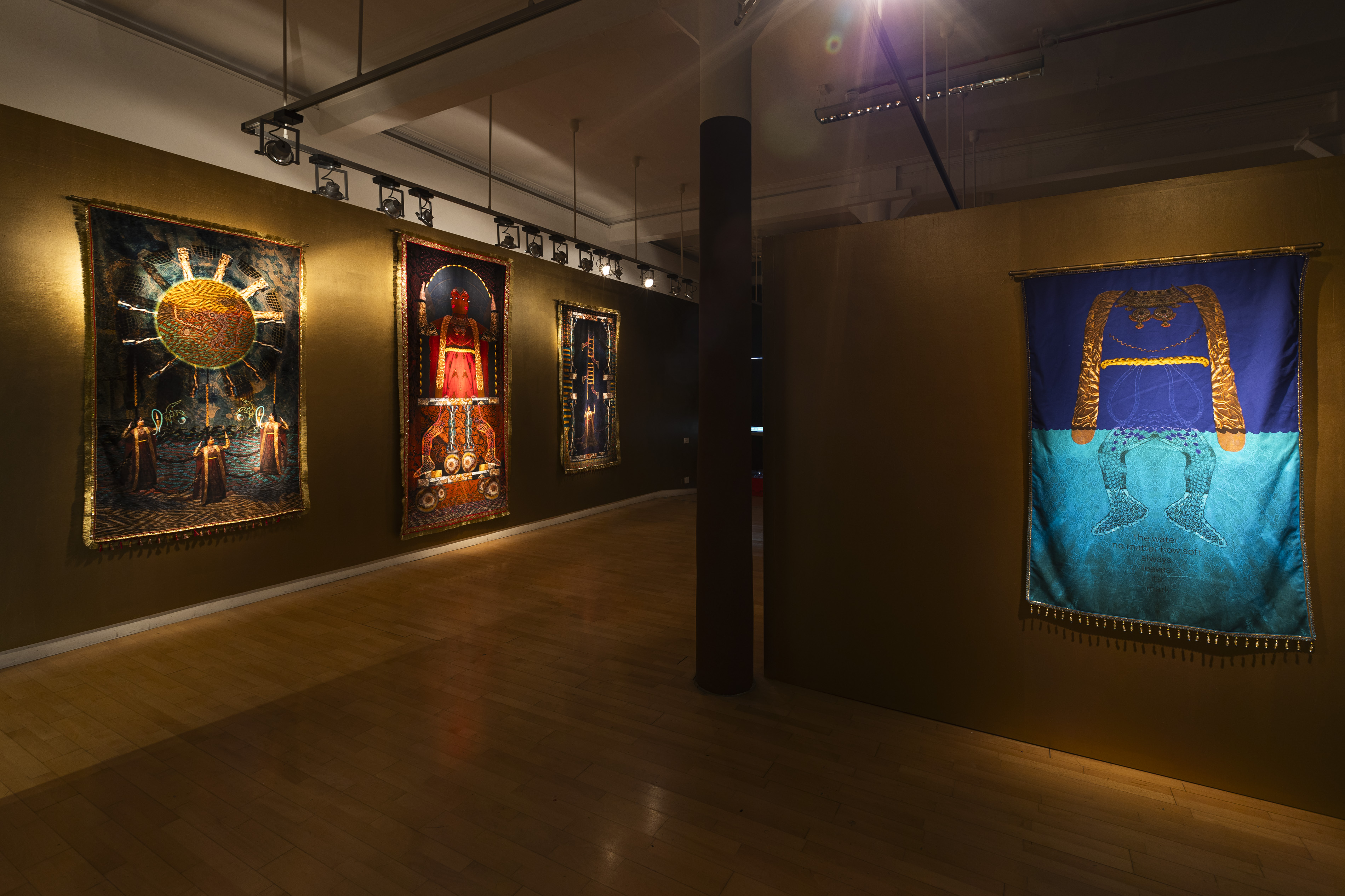 A gallery corridor at Streetlevel Photoworks hung with Aqsa Arif's tapestries, including a sun and robed figures, a red enthroned figure, a ladder motif, and a blue tapestry split between sky and water bearing the text "the water, no matter how soft, always leaves its mark