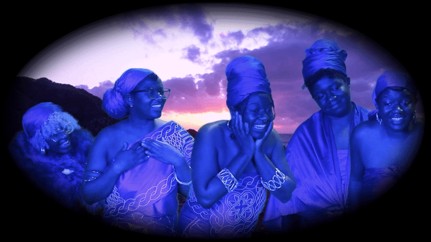 A group of women, all bathed in a blue light and wearing turbans and traditional dress, look excited.