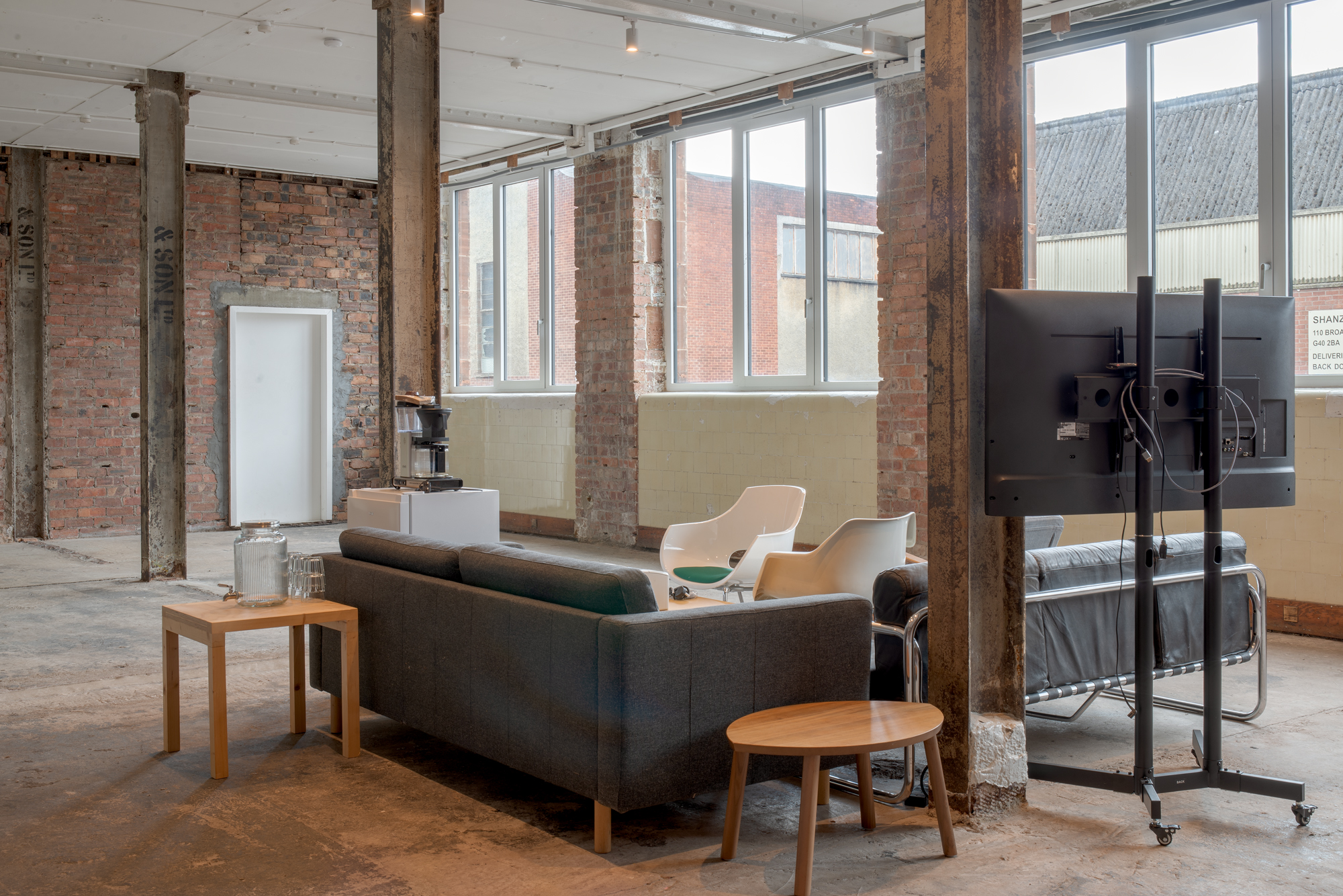 A furnished corner of a converted industrial space with exposed brick, steel columns and large windows, featuring a grey sofa, mismatched chairs, a drinks dispenser and a television on a mobile stand.