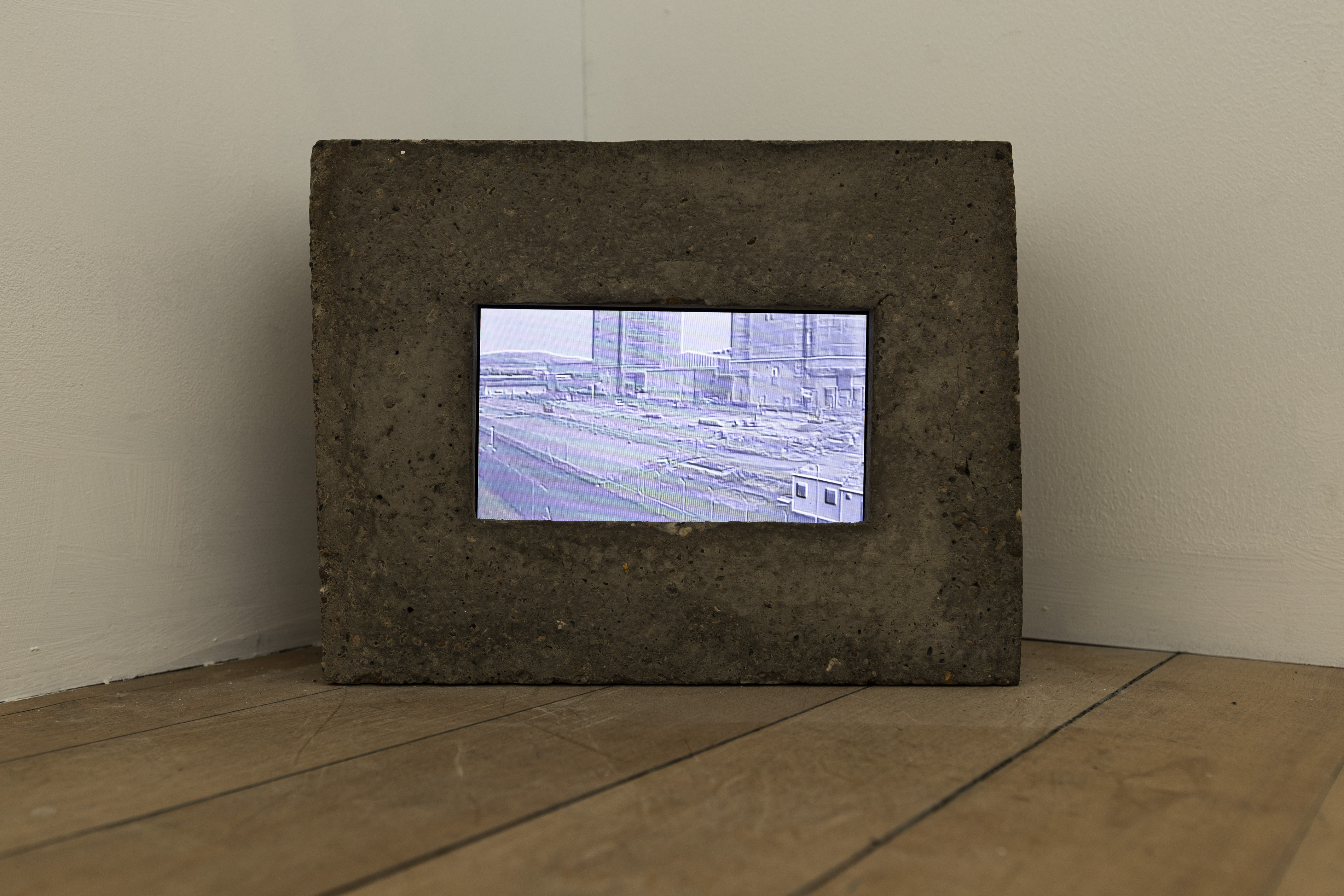 A small video monitor is embedded in a rough breeze-block or concrete frame, resting against a white wall on a wooden floor. The screen shows a washed-out, blue-toned surveillance-style image of an industrial or construction site, with tower-like structures, fencing, and a portable cabin visible.