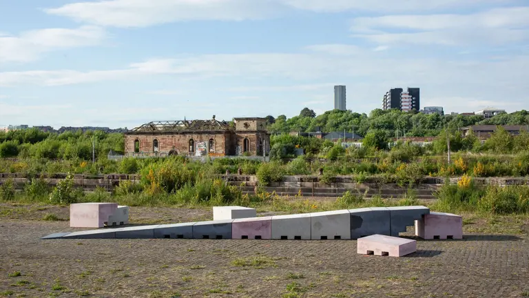 A series of abstract concrete blocks in grey and pink form a ramp. They are outside on cobbled ground with plants growing out of it.