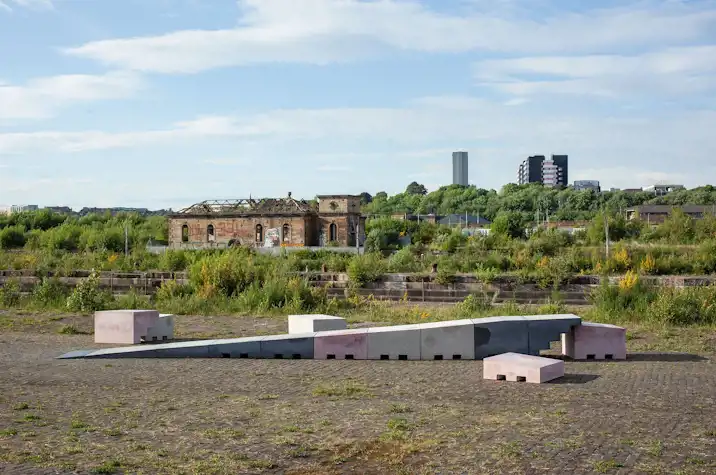 A series of abstract concrete blocks in grey and pink form a ramp. They are outside on cobbled ground with plants growing out of it.